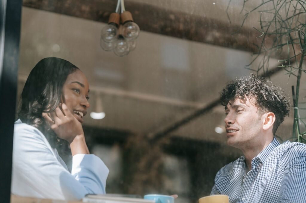 Twee volwassenen in gesprek aan een tafeltje bij een raam in een licht cafΓ©